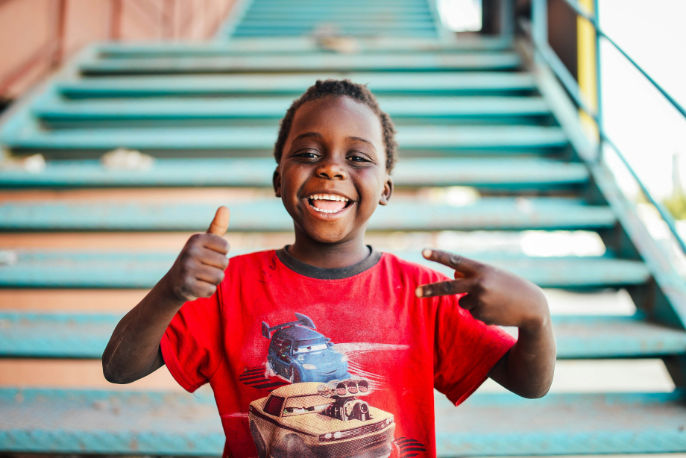 A young boy in red shirt pointing to his face.